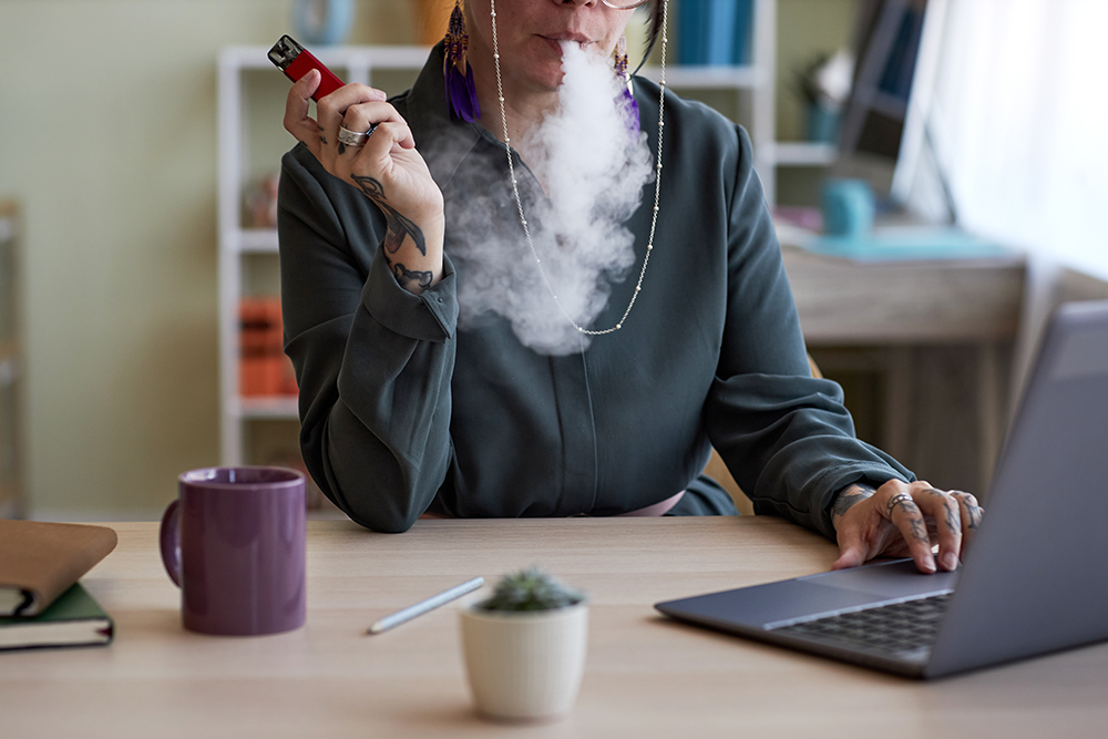 Close-up of of young creative female designer smoking electronic cigarette by workplace while sitting in front of laptop and networking
